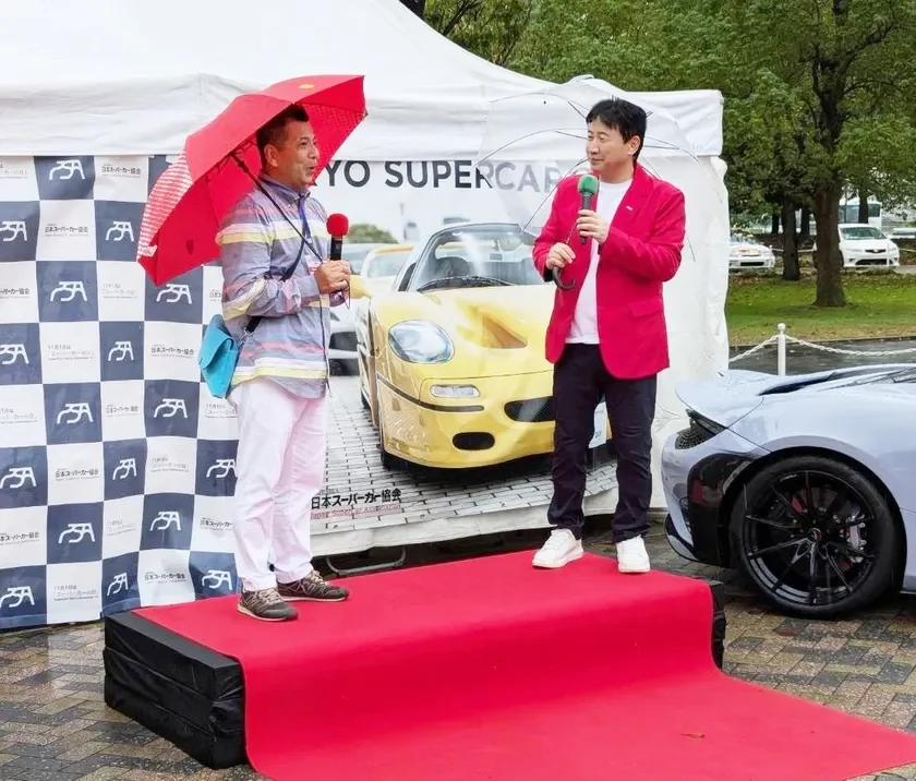 屋外のスーパーカーイベントで、二人の男性がマイクを持ち、傘を差しながらレッドカーペットのステージでプレゼンテーションを行っている