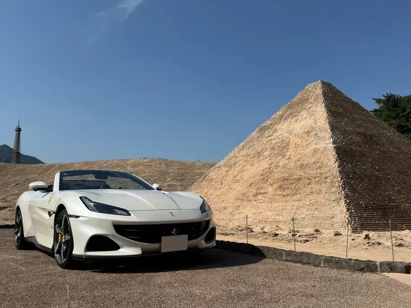 白いフェラーリのオープンカーが、巨大なピラミッドと遠くに見えるエッフェル塔のレプリカを背景に駐車されている風景