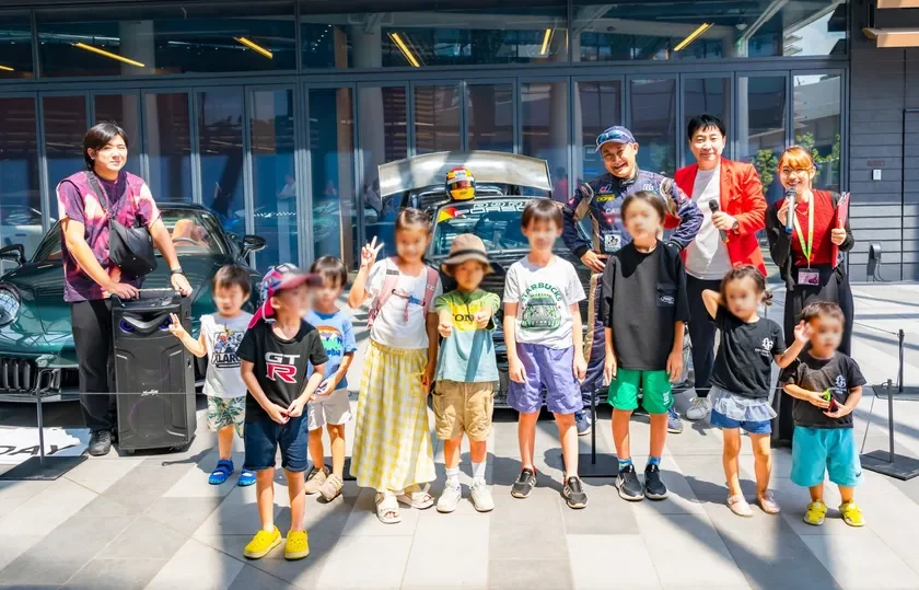 屋外で、レーシングカーとスポーツカーを背景に、子供たちと大人たちが笑顔で集合写真を撮っている様子