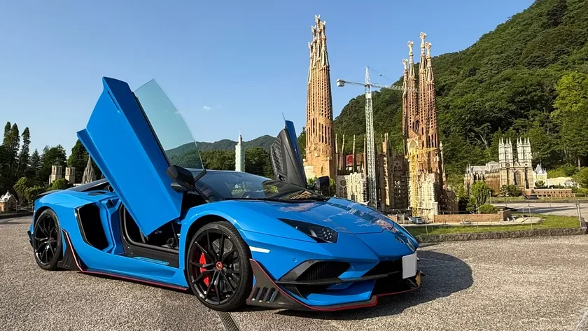 青いランボルギーニのスーパーカーが、サグラダファミリアなどのミニチュア建造物が立ち並ぶ屋外に駐車されている様子