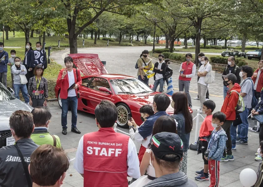 公園のような屋外で、赤いスポーツカー(フェラーリ)を囲んで多くの人々が集まっています