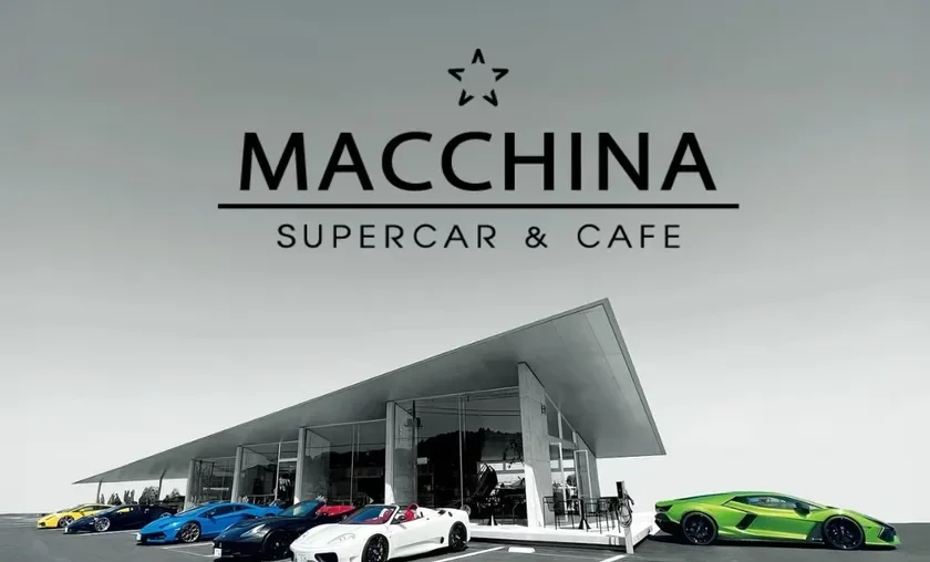 「MACCHINA SUPERCAR & CAFE」というロゴが上部に表示された、モダンな建物とカラフルな高級スポーツカーが並ぶ駐車場の風景